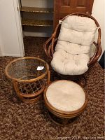 Full set of rattan swivel rocking chair, cushioned ottoman, and glass top end table on brown carpet.