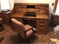 Front angle of oak roll top desk with matching upholstered office chair on casters in a carpeted room