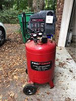 Full view of the red vertical air compressor with wheels and black handle, placed outdoors on a driveway with leaves on the ground.