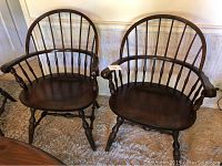 Two Windsor spindle arm chairs with curved backs, vertical spindles, and wooden armrests shown on carpeted room with wall paneling background.