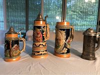 Five German beer steins displayed on a table with natural lighting and curtains in background, showing variation in size and design.