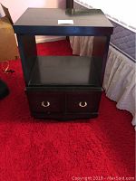 Front view of one black Curtis Brothers end table showing open shelf and two drawers with round brass handles on red carpet.