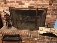 Photo showing brass three-panel fireplace screen in front of brick fireplace with fire grate inside and fireplace tool set on stand to the side. Log bin with faux birch logs on the hearth.