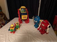 Six vintage toy robots displayed on a table with a black curtain background including a large red and yellow robot, two medium-sized blue and red robots, and three smaller colorful robots.