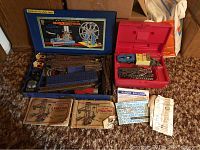 Two vintage Erector construction sets with metal parts, metal box, red plastic carrying case, and instruction manuals spread on carpet