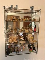 Full view of the metal and glass display shelf with three shelves featuring miniature collectible figurines including vintage Wade porcelain pieces and a Mickey Mouse figure. Shelf frame shows metal tarnish and bent front.