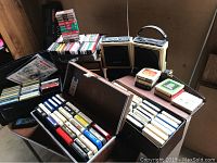 Wide angle photo showing two cases full of 8-track tapes and cassette tapes laid out on a table with two portable cassette players.