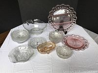 Overview of all 14 glassware pieces including various clear pressed glass bowls and plates and two pink glass plates with looped edges.