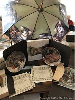 Image showing 6 collector plates from Edwin M. Knowles Amy Brachenbury Cat Tales Collection, each in original box with certificates and pamphlets, and a large cat-themed umbrella in the background.