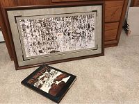 Framed artwork and book shown on carpet next to wooden furniture, print visible under glass.