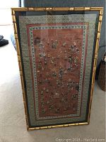 Full view of the framed embroidered silk panel showing intricate scenes of figures on a reddish-brown silk background with green and white patterned border and bamboo style gold frame.