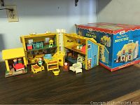 Photo showing open foldable yellow playhouse with multiple rooms, original box at right, and wooden peg people and furniture outside house
