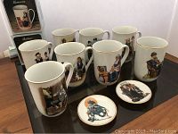 Photo of eight Norman Rockwell mugs and two small matching plates displayed on a table; two mugs shown with original boxes stacked behind.