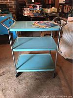 Full view of the blue metal cart showing three shelves and chrome handles with magazines on top