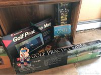 Wide shot showing boxed golf practice mat, golf ball shagger, World Atlas of Golf book, and partially visible golf practice net.
