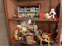Photo showing two shelves of various vintage children's toys including a metal doll house, doll house furniture pieces, a white snowman toy figure, the Samsonite Ark with original box, and a bucket with small toys.