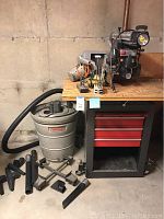 Radial arm saw on table with rolling tool cabinet, shop vac and multiple tool accessories on and around it.
