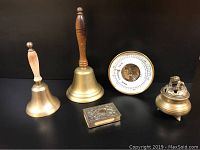 Photo of two vintage brass bells with wooden handles, brass barometer, Chinese brass incense burner with Fudog dog, and engraved brass match case on black background.