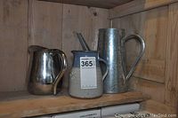 Three jugs on a wooden shelf: stainless steel jug, clay jug, and tall tin water can