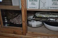 Placements, bowls, gold tone wire basket, and tablecloth sets visible in wooden cabinet shelf
