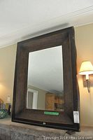 Framed mirror with dark brown, textured wooden frame showing the entire mirror and frame against a wall with lamp reflected and some damage visible on frame corners.
