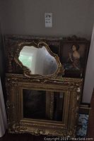 Photo showing multiple large ornate gilded vintage frames and a uniquely shaped mirror leaning against a wall, with a framed portrait of a woman