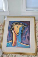 Framed colorful abstract portrait painting on a floral couch with white window frames in the background.
