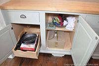 Image shows open cabinet with upper shelf containing wooden bowl with utensils and cloth, sealed cardboard boxes below, partially opened drawers and doors.