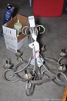 Metal hanging light fixture shown from above with six candle-style bulb holders and decorative curved metal arms. The fixture is placed on a concrete floor inside a storage setting.