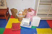View of collection of wicker baskets, pink fabric basket with blanket, plastic storage bins including a white bin with lid, and small red woven basket placed on colorful foam mat flooring.