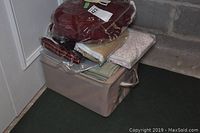 Box containing assorted folded drapes and fabric pieces stacked inside and on top of a soft storage box placed on floor next to wall.