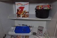 Photo of shelf showing a boxed tomato strainer canning tool and black canning pot with lid, white shelf interior.