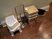 Photo showing white plastic step stools with blue nonslip dots, a vertical metal toilet tissue holder, small beige plastic bins, metal trash bin, and woven baskets on hardwood floor.