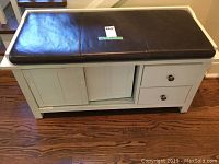 Front view of white wooden bench showing black cushion seat, two small drawers on right, sliding cupboard doors in center, wooden paneled design, scratches and wear visible