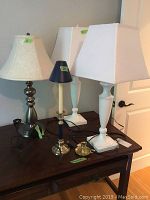 Front view of five items on a dark table, showing two white lamps, a metal based lamp, and three candle holders.