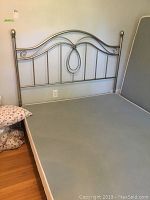 Metal queen size bed frame with silver decorative headboard shown against wall with adjacent bedding pillows.