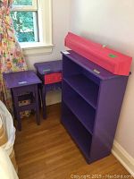 Purple bookshelf with two shelves against the wall and two smaller tables stacked beside it: one small square purple table and one purple side table with pink drawer accents. The furniture is painted wood, designed for a girl's room.