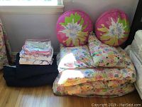 Two sets of folded girls single bed linens stacked with two round pillows on top featuring pink and green floral designs, along with a white duvet underneath and dark blue comforter beside them