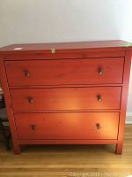 Front view of red IKEA Hennes dresser with three drawers and black round knobs, showing scratches and wear.
