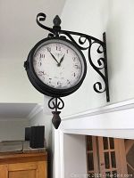 Side view of the black metal double-sided wall clock mounted on white wall with decorative curled wrought iron bracket, showing clock face with Paris motif.