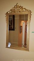 Full view of gilded ornate framed mirror hanging on a wall, showing its overall rectangular shape and decorative top.