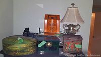 Photo showing five items including a painted oval wooden box with nature-themed painting, a green mirrored jewelry box, a tin container with ornate design and color, and a silver-tone loving cup style table lamp with fabric shade