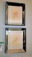 Photo of two beveled mirror framed prints hung vertically on a wall, showing reflective wide silver frames and the detailed sketches inside.