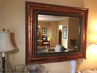 Full view of mirror mounted on wall above furniture, showing rectangular shape, beveled mirror edges, and copper-tone resin frame with textured finish and decorative detail.