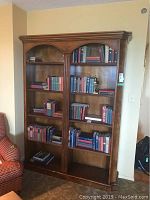 Full view of wooden bookshelf holding multiple classic hardcover books, organized mostly by size and color, located against wall beside a red upholstered chair.