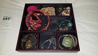 Top-down photo showing a divided jewelry box with assorted necklaces, bracelets, faux pearls, earrings, a watch, and a green compact mirror with floral decoration
