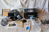 Garmin StreetPilot III GPS unit with accessories including manual, charger, and mount; Motorola Razr and Samsung Mantra flip phones with chargers and accessories laid out on cloth background