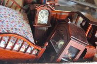 Close-up photo showing mini wooden bed with floral mattress, small grandfather clock and glass-front cabinet with decorative items inside