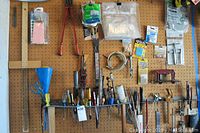 Overview of multiple hand tools on wooden pegboard, including screwdrivers, pliers, scissors, funnel, files, and packaged hardware