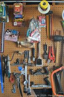 Pegboard with assortment of hand tools including saws, hammers, wrenches, scissors, pliers, brushes, small clamps, and packaging with small hardware items.